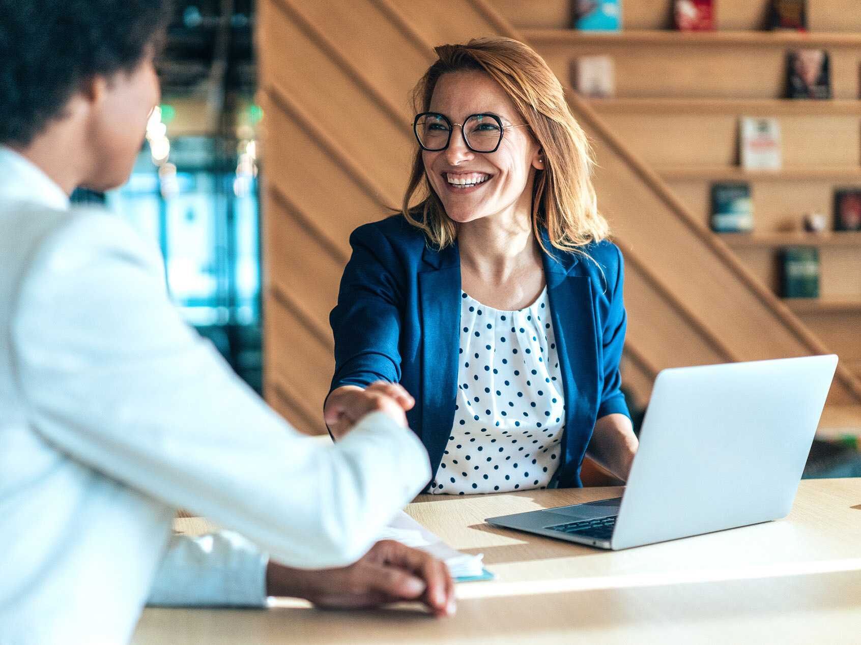 Two professionals shaking hands across a desk with a laptop during a workplace meeting