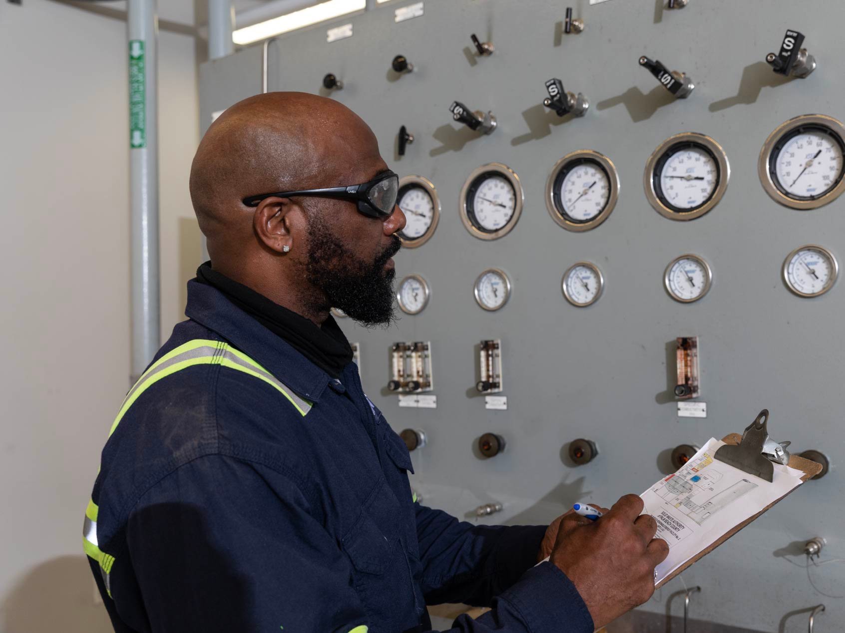Plant operator in safety gear recording readings on a clipboard beside a panel of pressure gauges