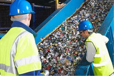 Workers inspect aluminum cans moving down a metal chute in a recycling facility.