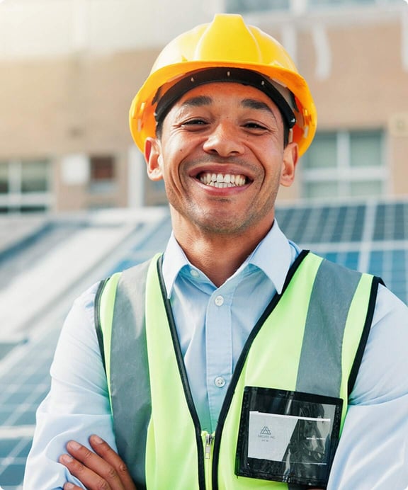 Smiling worker in a yellow hard hat and safety vest standing outdoors at a facility